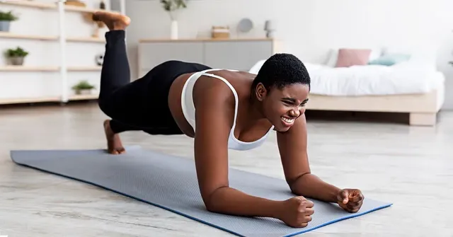 Woman in black leggings and white sports bra doing a plank exercise on a blue yoga mat in a bright bedroom.